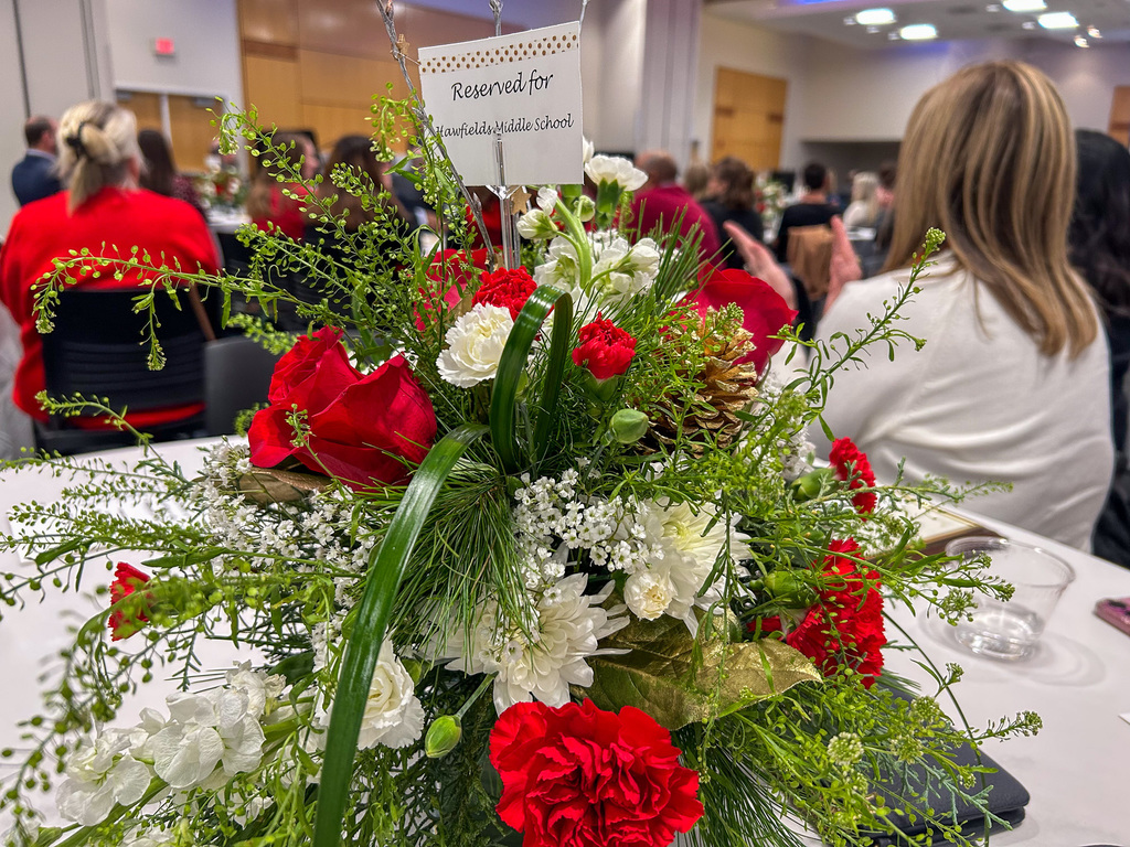 A close-up shot of a festive centerpiece featuring red roses, white flowers, and greenery. A card on a stand above the centerpiece reads, "Reserved for Hawfields Middle School." Attendees are seated in the background.
