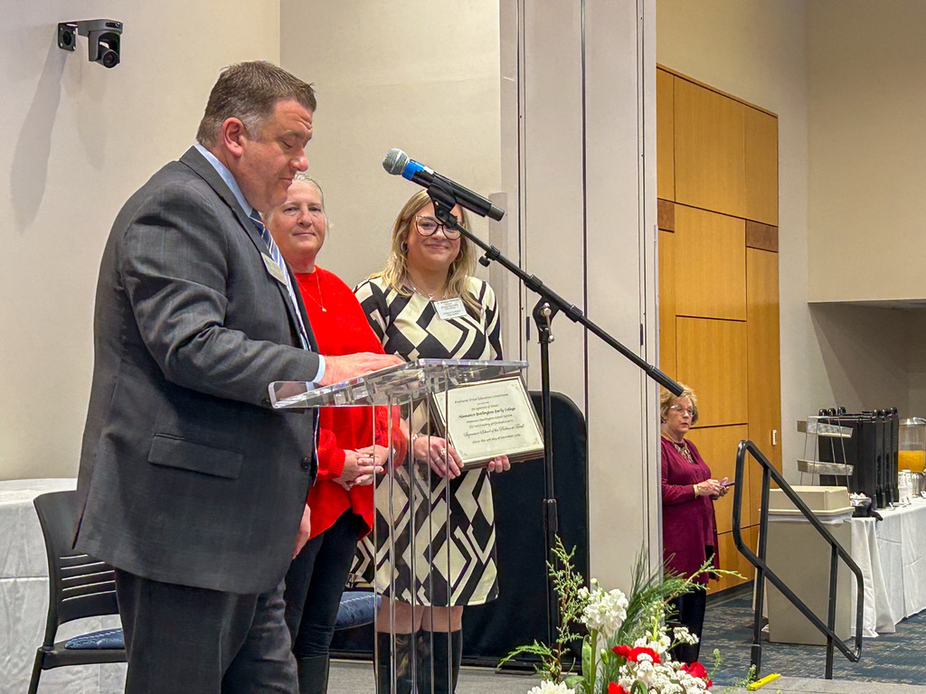 A man in a suit stands behind a lectern and speaks into a microphone while presenting a framed certificate to two women standing beside him on a stage.