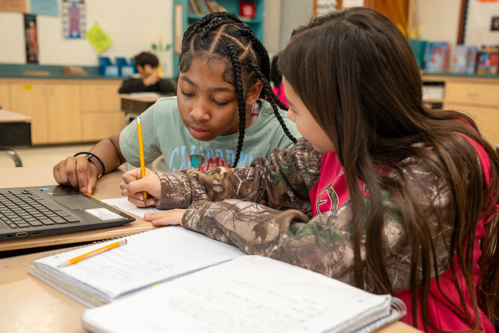 Two girls work collaboratively during science time, writing in a journal in front of a Chromebook