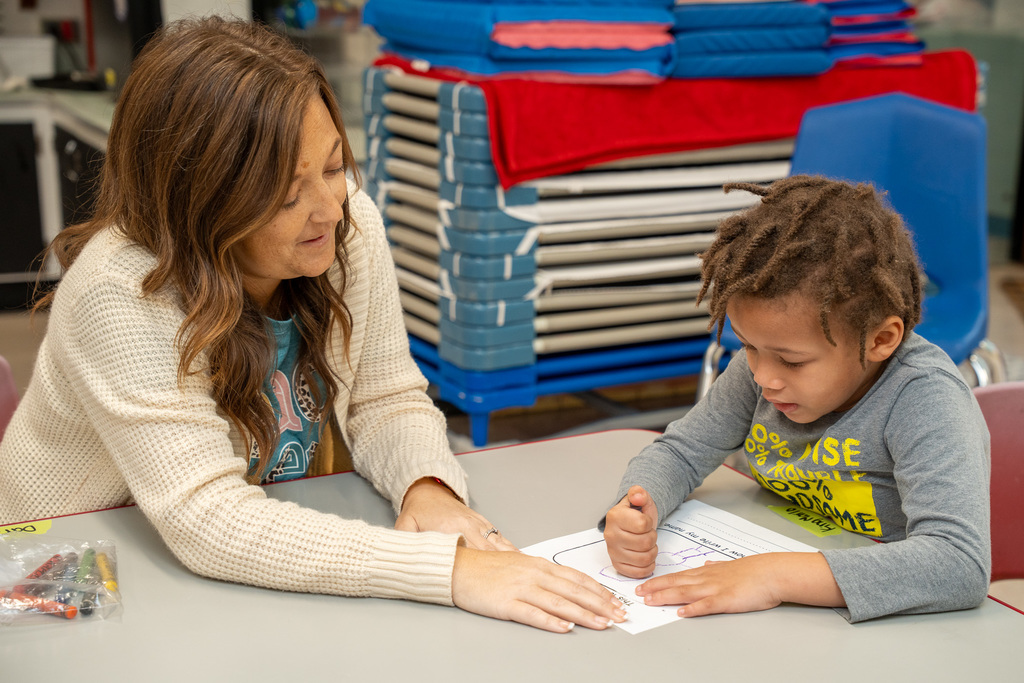 A Pre-K teacher works closely with a student to draw a picture on paper