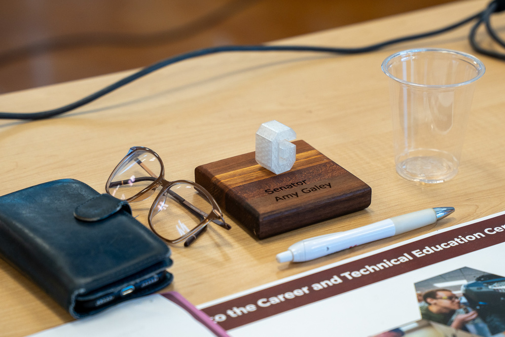 A carved nameplate for Senator Amy Galey with a 3D-printed G on top.  Around it are a clear cup pen, glasses, and a phone with a leather case.