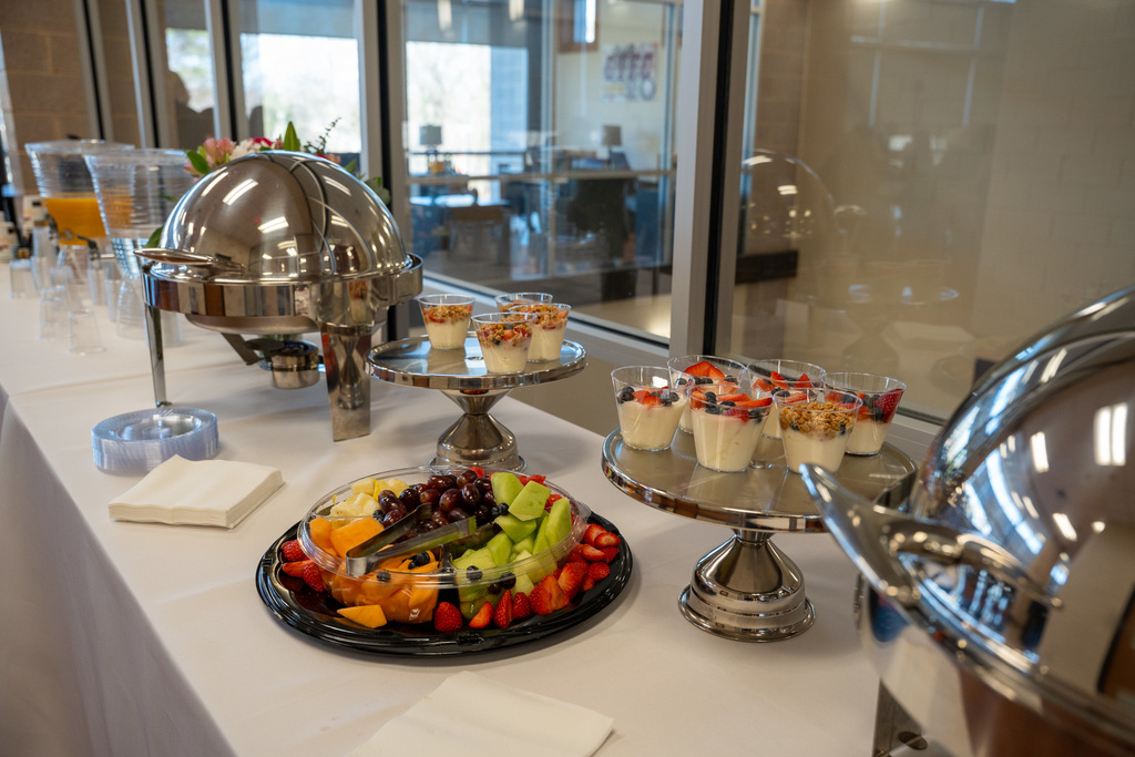 A breakfast spread with yogurt parfaits, fresh fruit, covered silver dishes, napkins, and drink options.