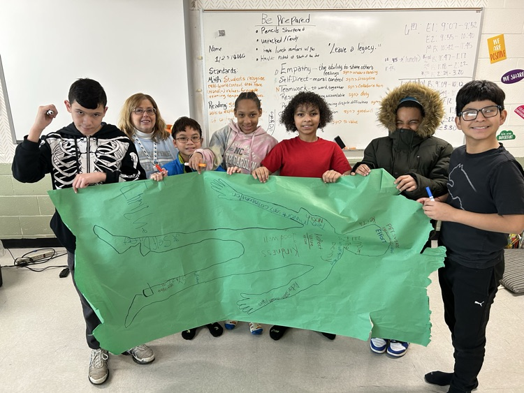 a small group of students holding a green poster with an outline of a person and inside or written various vocabulary words related to empathy