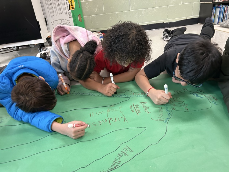 A smaller students on the floor gathered around a green poster writing vocabulary rewards related to empathy inside the outline of a person