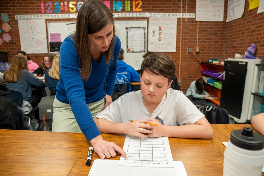 A teacher leans over a student and points to a two-step math problem