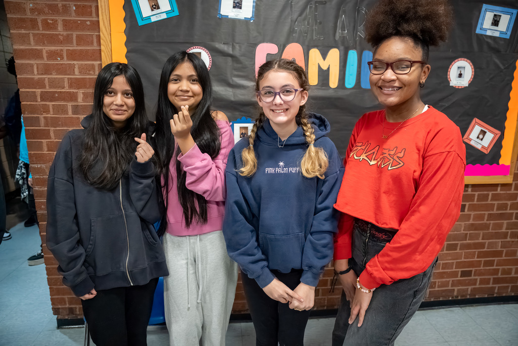 Four students pose in the hallway in front of a black bulletin board and brick wall