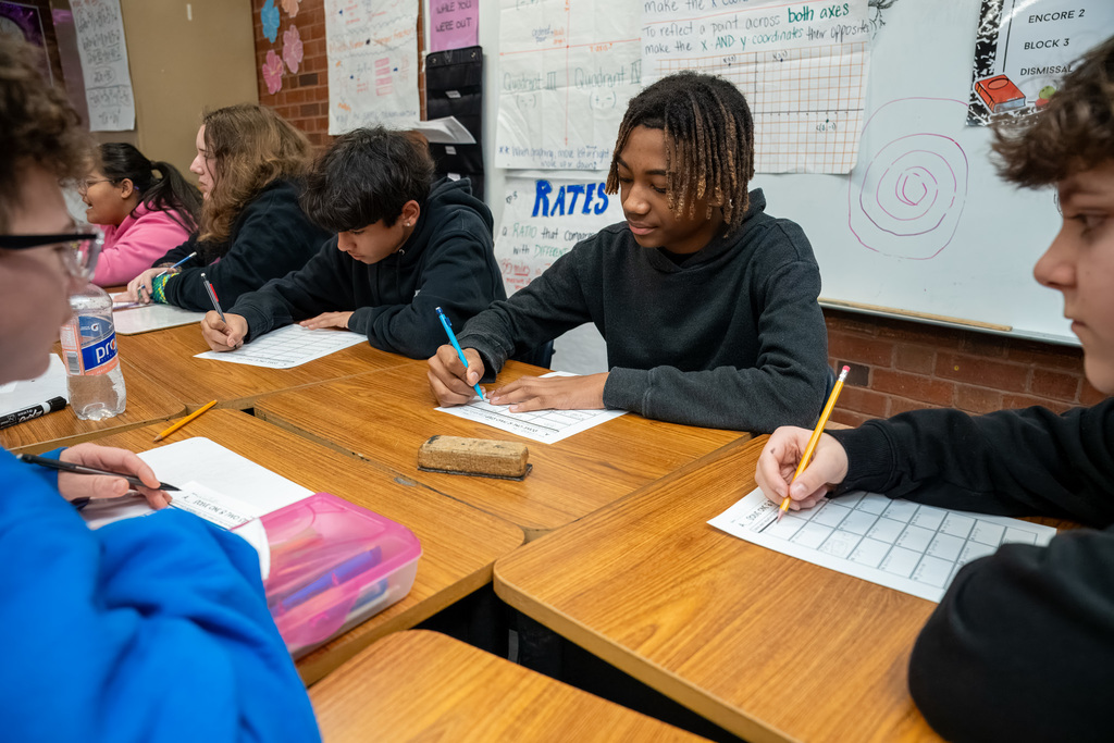 Multiple middle school students at their desks which are clustered together, all working on two-step math problems