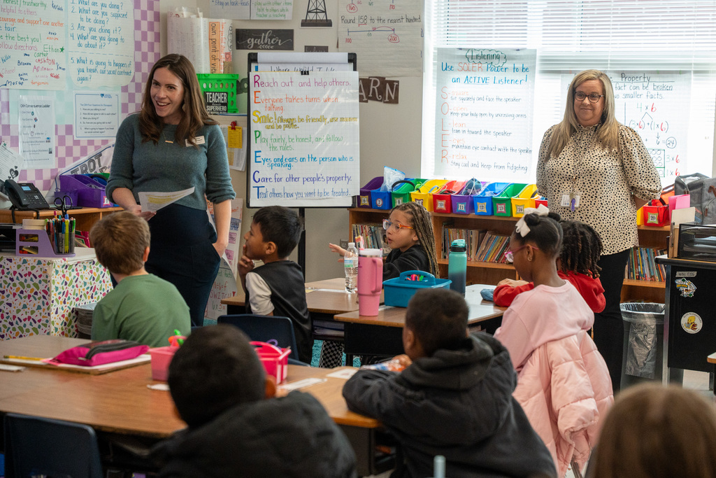 Two women present to a classroom of third-grade students at Elon Elementary. One woman in a teal sweater gestures while speaking, and another in a leopard print top stands nearby. Students are seated at desks listening attentively, with classroom posters and the "RESPECT" acronym visible on the wall behind them.