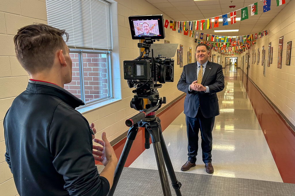 Dr. Fleming being filmed in a school hallway decorated with international flags. A videographer operates a professional camera on a tripod as Dr. Fleming speaks to the camera in a suit and tie.