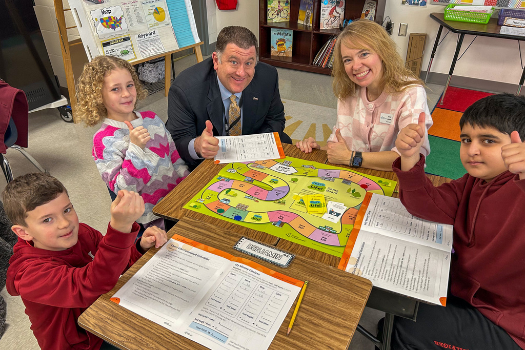Dr. Fleming sits at a table with three third-grade students and a Junior Achievement volunteer, all giving thumbs up while playing the "That's Life!" financial literacy board game.