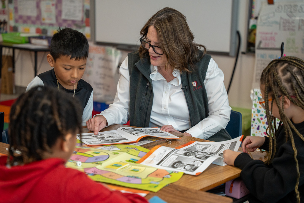 President Connie Book of Elon University sits at a table with third-grade students, reviewing Junior Achievement financial literacy materials including colorful game boards and worksheets.