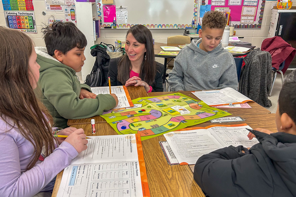 Elon Elementary principal Cara Hurdle works with a small group of third-graders at a table, guiding them through the "That's Life!" game as they make decisions about earning, saving, and spending money.