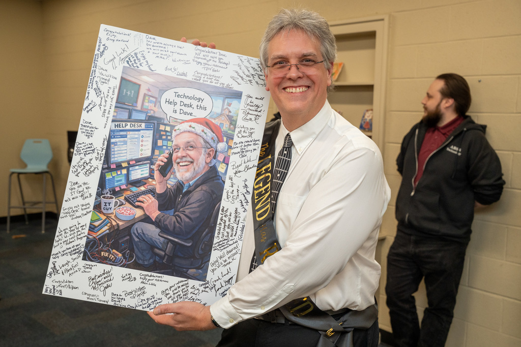 Dave holding a signed photo caricature of him answering help calls at a computer desk