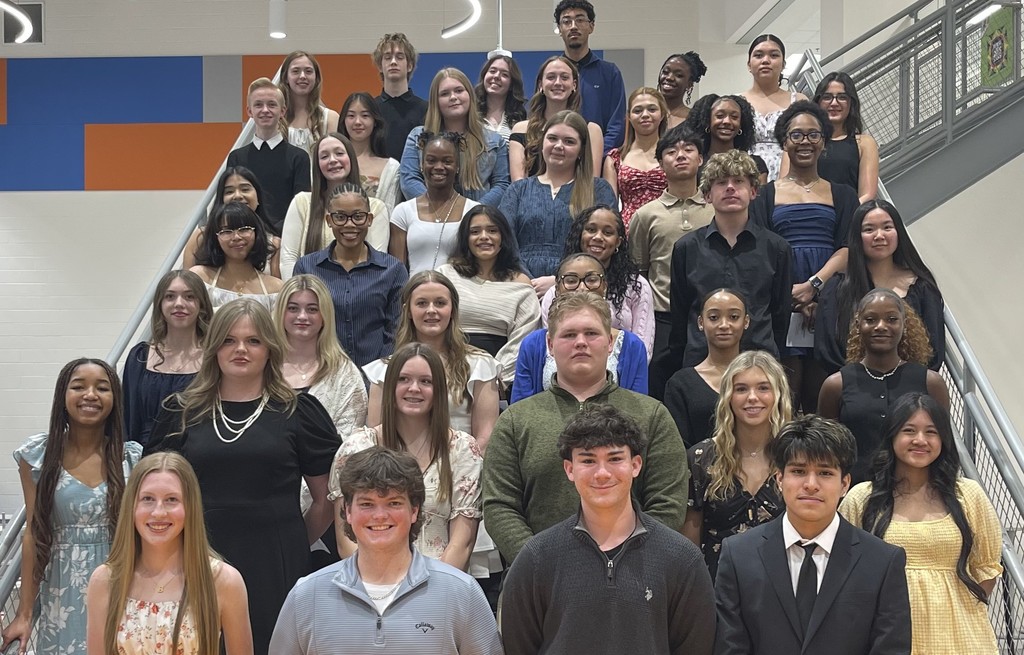 A large group of students, standing in organized rows on a staircase, smiling in a posed photo, before the National Honor Society Induction Ceremony begins