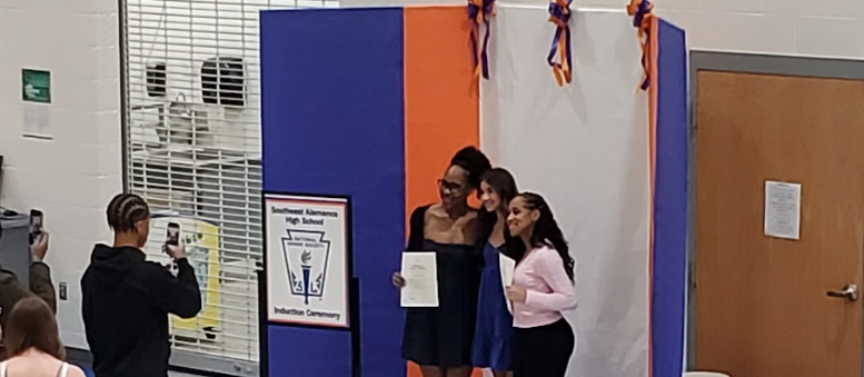 three students hold their NHS membership certificates and stand next to an NHS sign, smiling for the camera as another student takes their photo