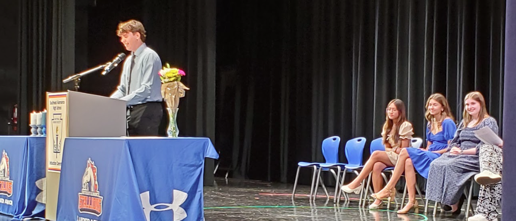 NHS officers sit in chairs while a male student stands at the podium introducing new members to the NHS