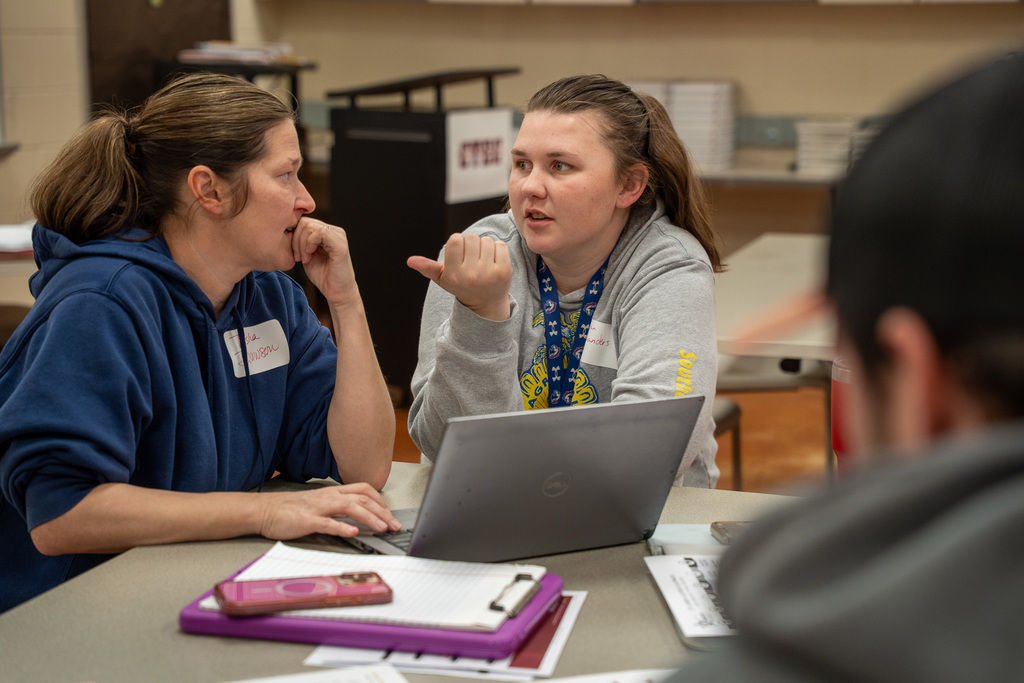 Two female teachers sit side by side at a table in close conversation during a professional development session. One teacher gestures with her hand as she speaks while the other listens attentively. Both have name tags and laptops open in front of them.