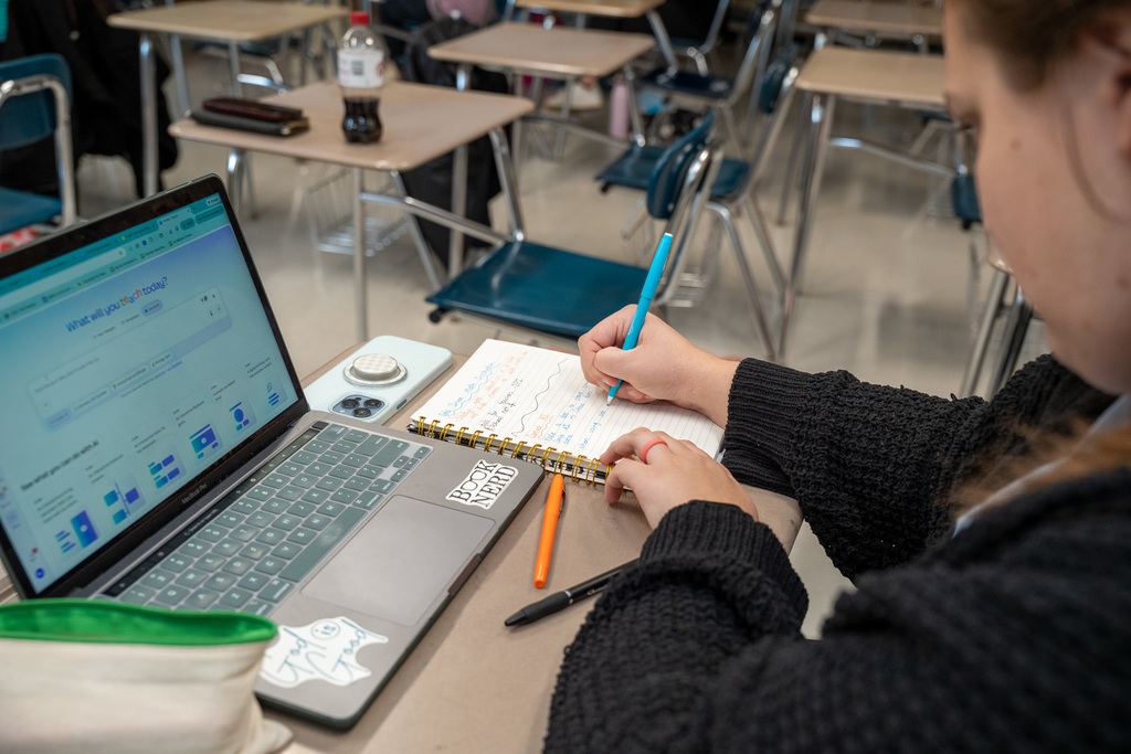 A close-up of a teacher's hands writing notes in a spiral notebook with a blue pen. An open laptop displaying an educational planning website is positioned to the left. A sticker on the laptop reads "Book Nerd."