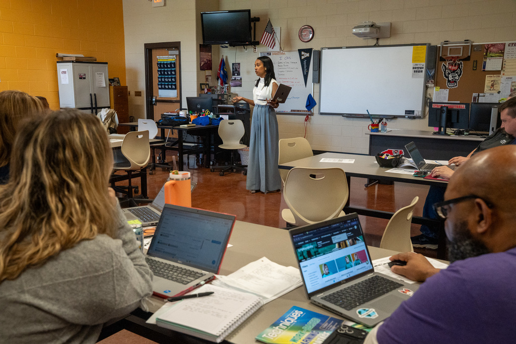 A female presenter stands at the front of a CTE classroom holding a laptop and addressing a small group of seated teachers. A whiteboard behind her displays an "I CAN" learning objective. Teachers in the foreground have laptops and notebooks open on their desks.