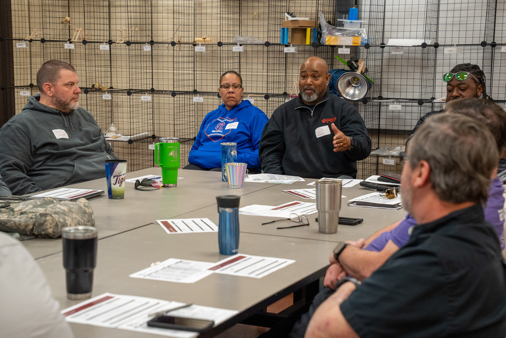 A group of CTE teachers and business advisory members sit around a large table engaged in discussion. A man in a black jacket gestures as he speaks while others around the table listen. Name tags and printed materials are visible on the table. The setting appears to be a workshop or lab classroom with storage shelving in the background.