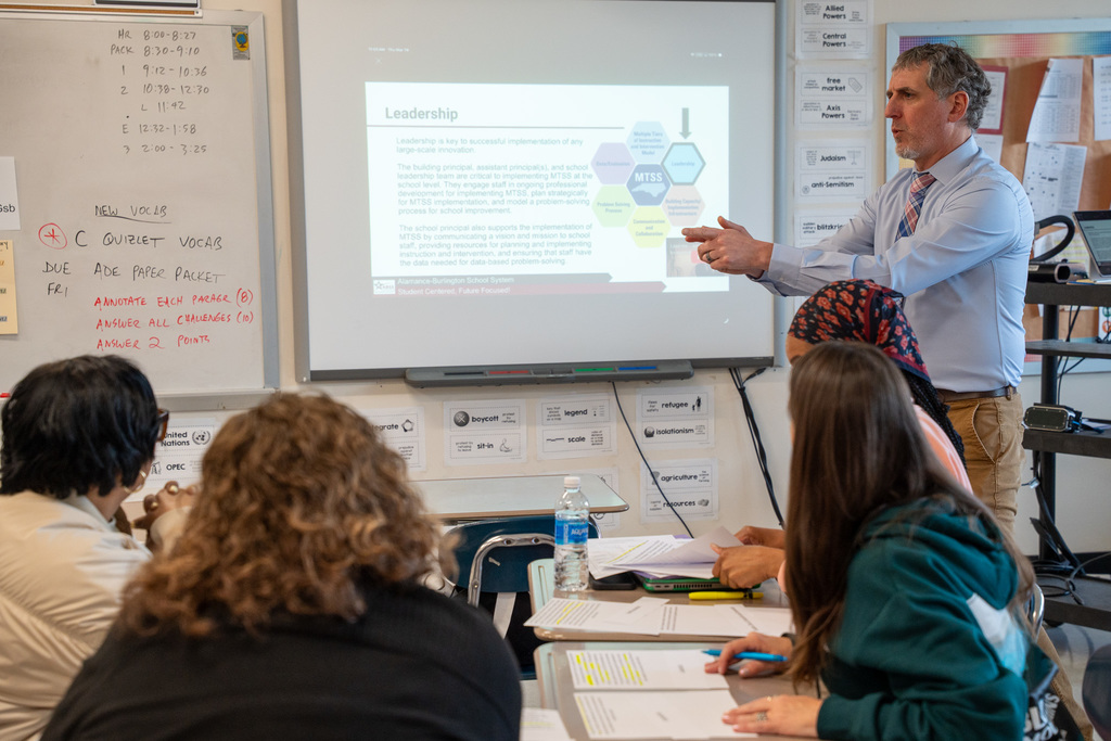 A male presenter stands at the front of a classroom pointing to a projected slideshow with a slide titled "Leadership." Several teachers are seated at desks in the foreground with papers and water bottles, facing the presenter attentively.