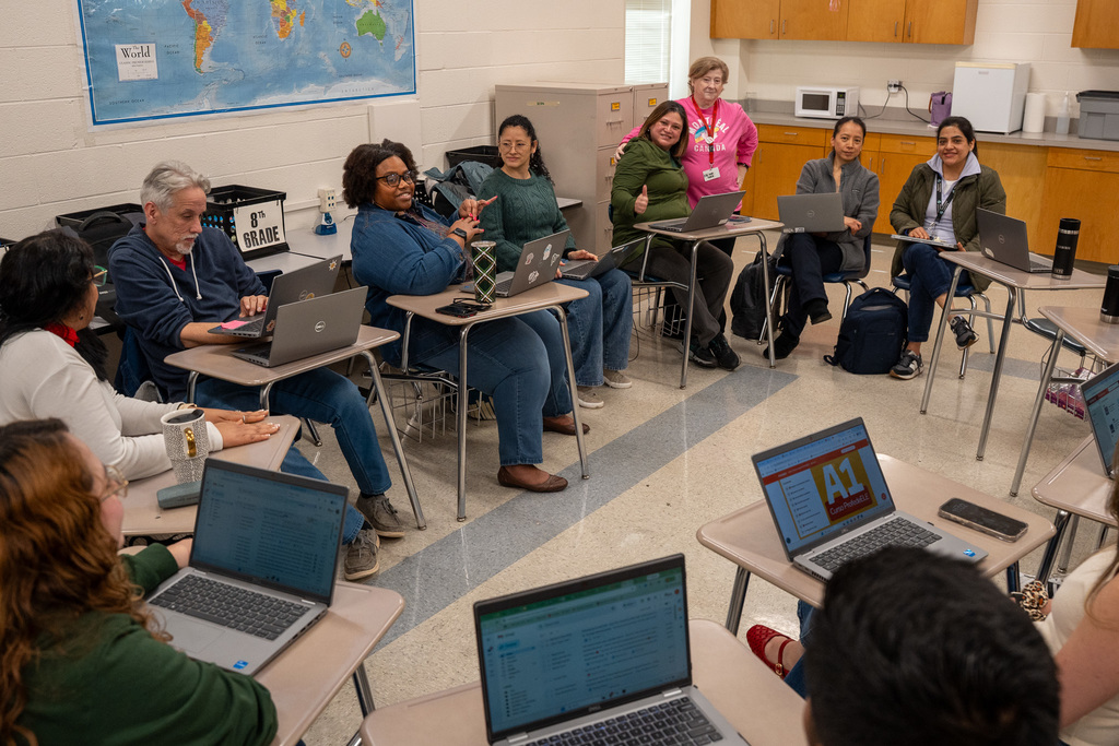 A large group of teachers sit in a circle arrangement at individual desks in a classroom, each with a laptop open. A world map is visible on the wall in the background. Several teachers in the background smile and give a thumbs up toward the camera.