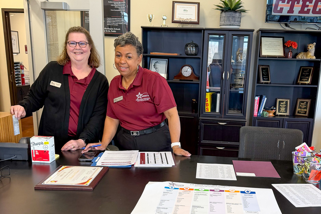 Two women wearing maroon ABSS CTE polo shirts stand together smiling behind a reception desk at the ABSS Career and Technical Education Center. The desk holds printed materials including a CTE Career Clusters and Pathways brochure, certificates, and registration paperwork. Award plaques and trophies are displayed on shelving in the background.