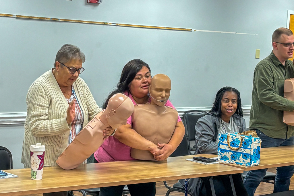 A Pre-K teacher in a pink shirt practices the Heimlich maneuver on a CPR training mannequin during a First Aid recertification session. An older woman in a white cardigan stands to the left offering guidance. A woman in a gray hoodie is seated at the table in the background, and a man in a green shirt stands to the right holding another training mannequin.