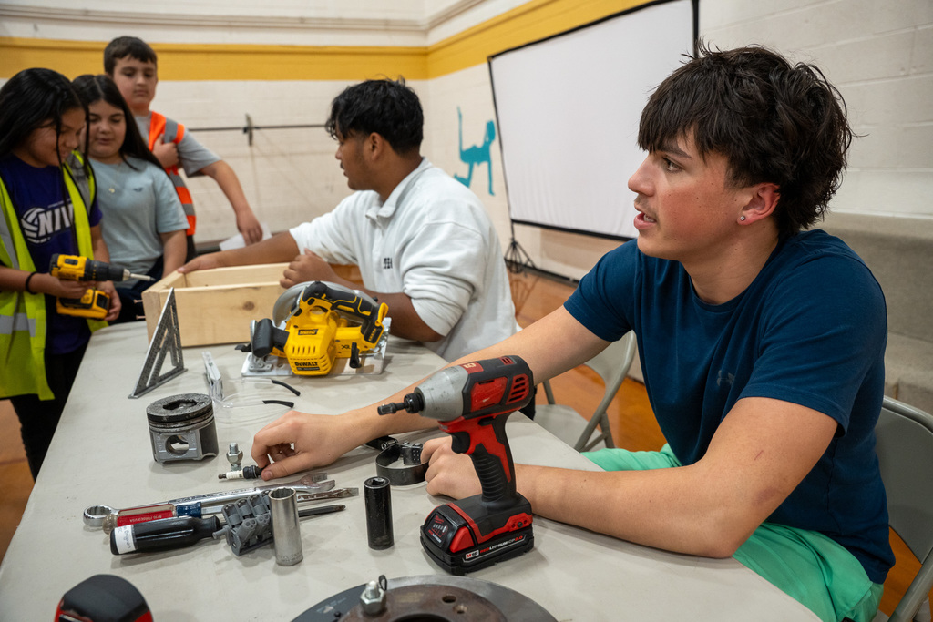 A high school student sits at a table displaying a mix of Automotive and Carpentry tools, including a circular saw and an engine piston. He explains the different mechanical career paths to elementary students wearing yellow safety vests.