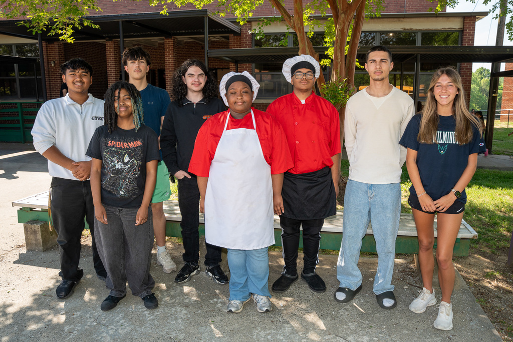 A group portrait of seven high school students from the Career & Technical Education Center standing outdoors at Haw River Elementary. The students represent various career paths, including two in culinary uniforms (red chef coats and white aprons/toques), one in a nursing fundamentals shirt, and others in casual attire representing automotive, carpentry, and animation programs.