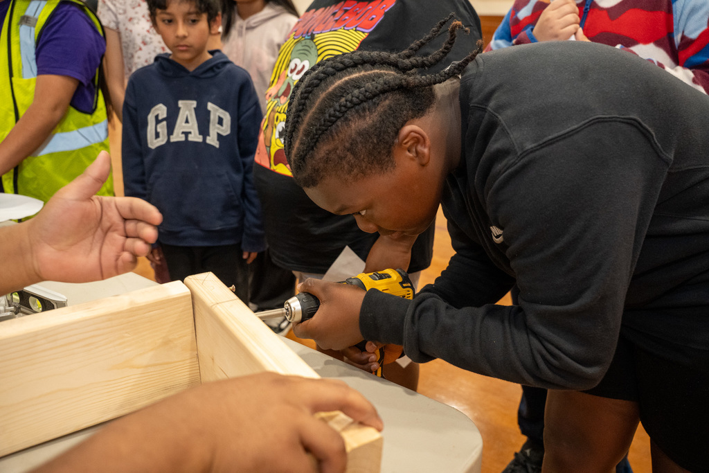 Close-up of an elementary student focused intently on using a power drill to drive a screw into a wooden frame during a Carpentry demonstration, while a high school mentor provides guidance.