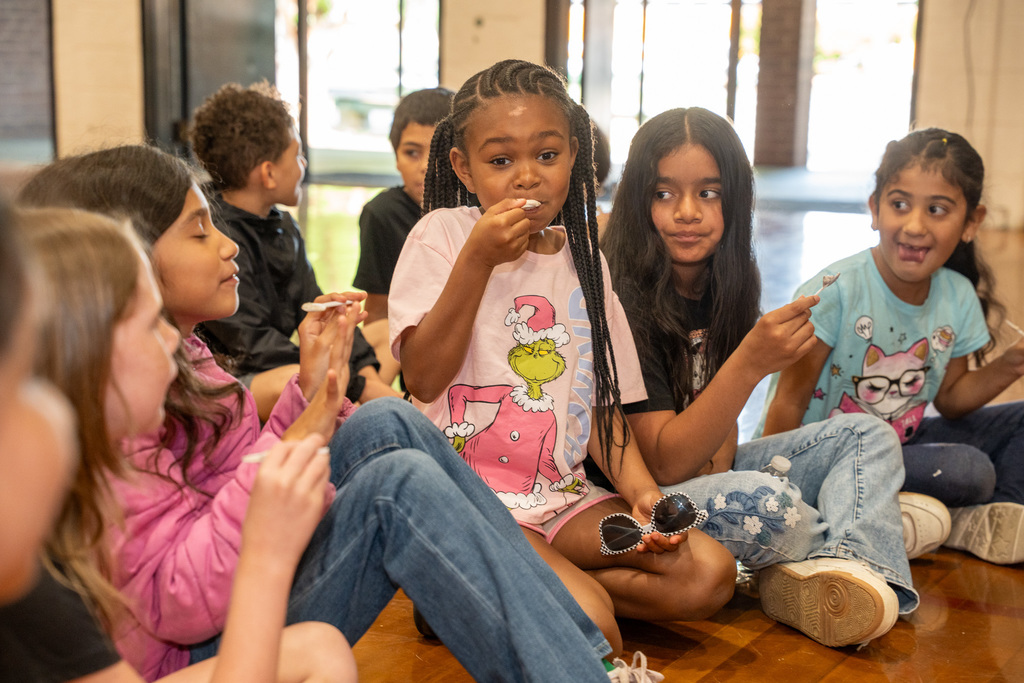 A group of elementary school children sit on a wooden floor, happily tasting samples provided by the culinary arts program. A girl in the center wearing a pink Grinch shirt enjoys a small spoonful of food.