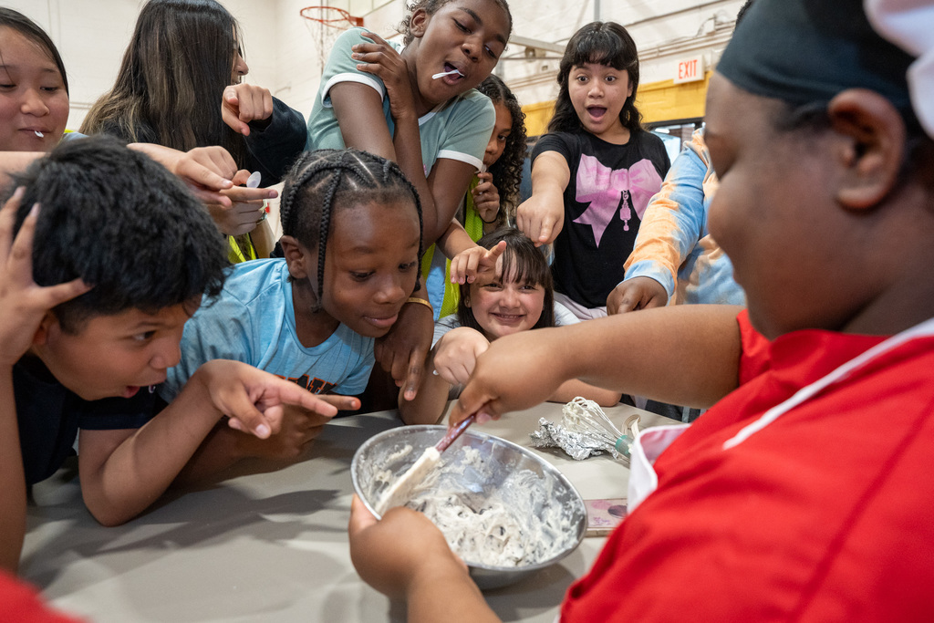 A group of elementary students lean in with excitement, pointing and cheering as a high school culinary student in a red chef’s coat and white apron whisks a bowl of frosting or batter.