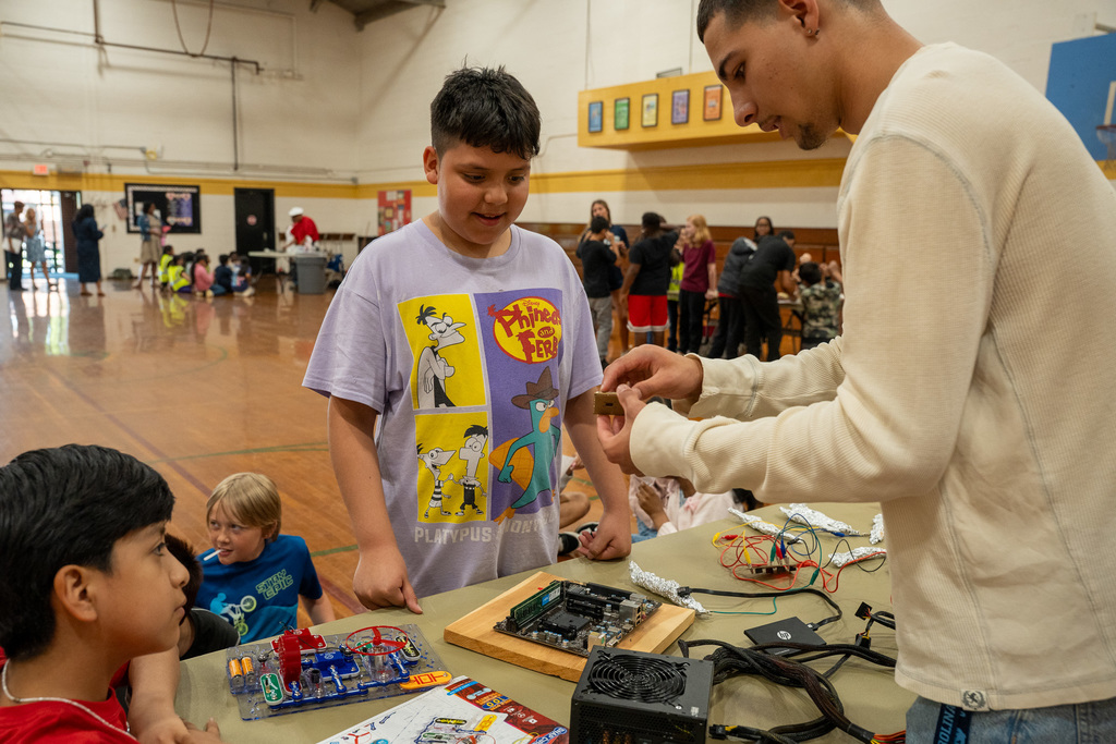 A high school Computer Engineering student shows a small electronic component to an elementary student. The table is spread with hardware including a motherboard, a power supply unit, and a Snap Circuits kit for hands-on learning.