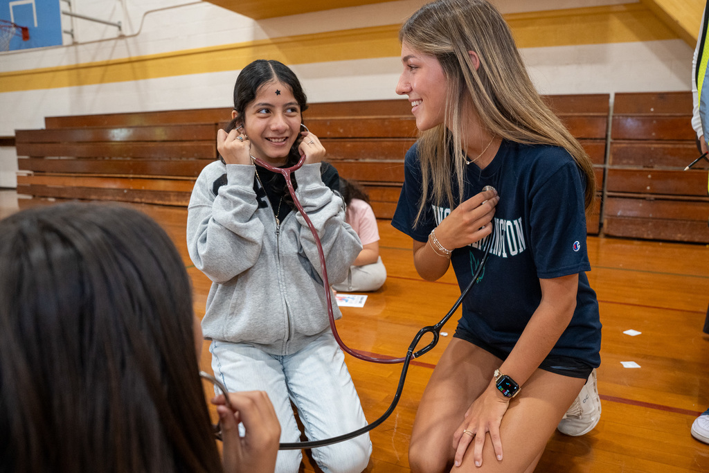 A high school nursing student smiles as she helps an elementary school girl use a stethoscope. The younger student, sitting on a gym floor, holds the earpieces to her ears with a look of curious wonder as she listens to a heartbeat.