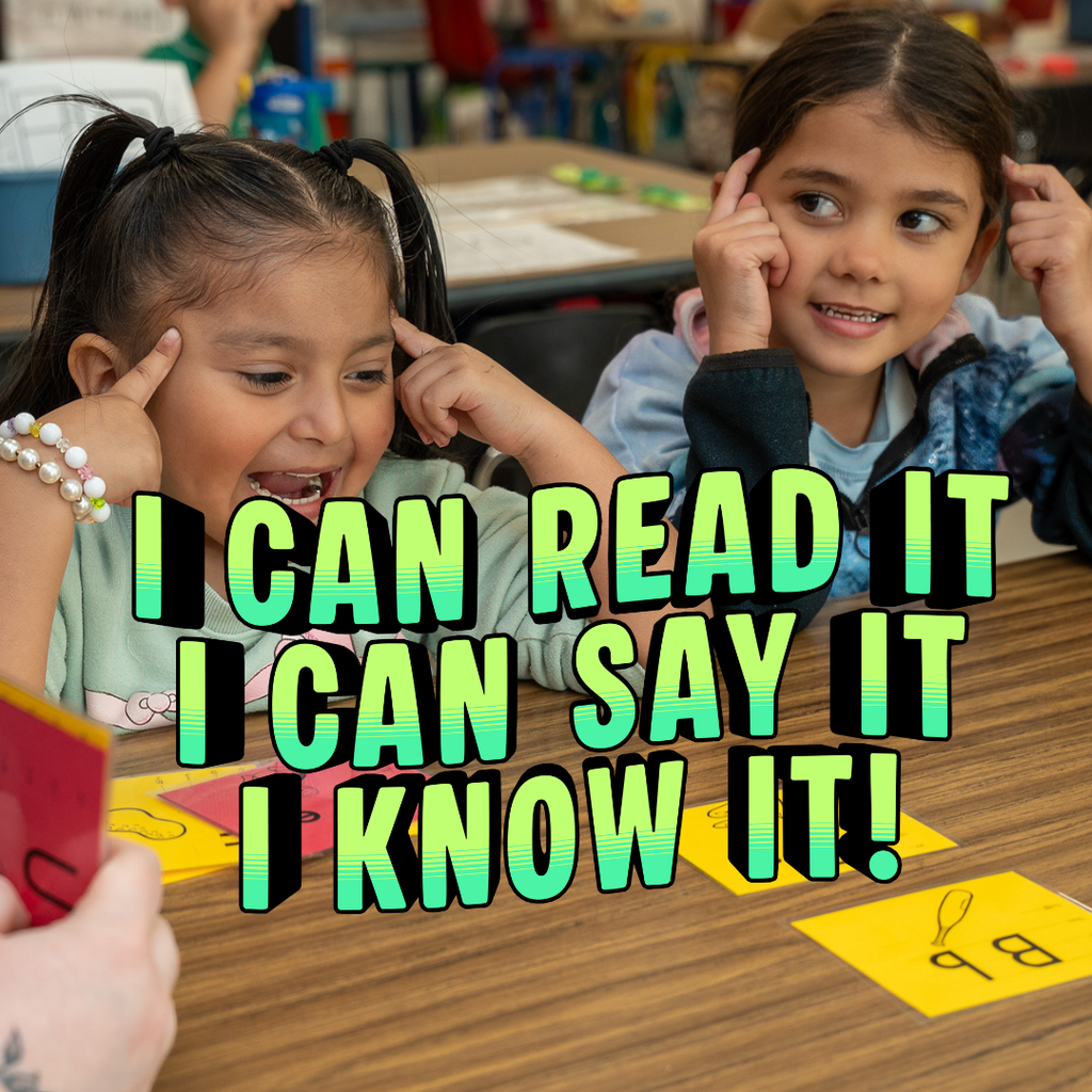 Two young girls sit at a classroom table, smiling and playfully pointing to their heads as they participate in a reading activity. In the foreground, flashcards with letters and drawings are visible. Bold, stylized text overlaid on the image reads, "I CAN READ IT, I CAN SAY IT, I CAN KNOW IT!"