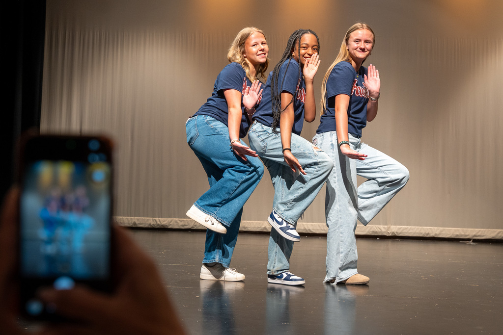 Three young female student-athletes in matching navy "Huskies" t-shirts and jeans strike a playful, coordinated pose on a stage, while a hand in the foreground holds up a smartphone to capture the moment.