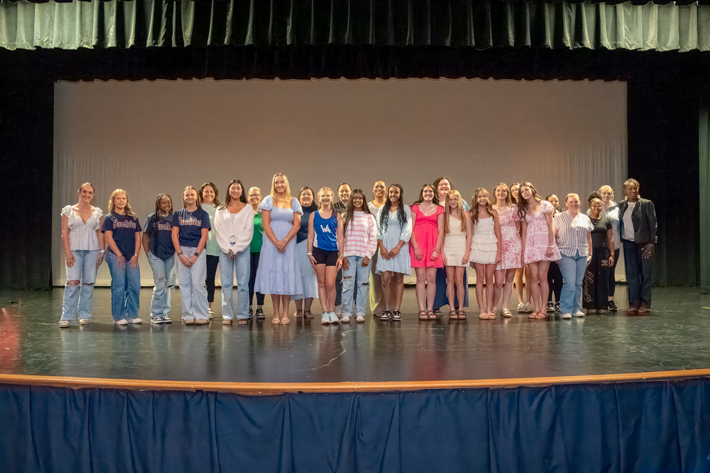 A full-stage view of the diverse group of female student-athletes and school leaders lined up horizontally on the stage, showcasing the significant growth in participation within the district.
