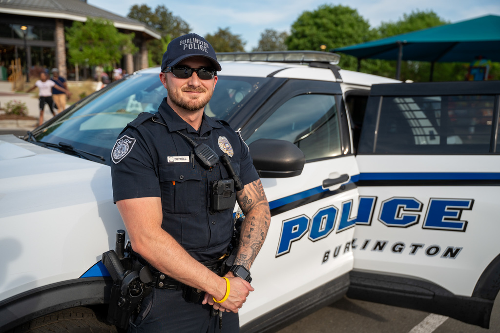 A Burlington Police officer in uniform stands proudly in front of a white and blue patrol vehicle at the park, representing the first responders participating in the event.