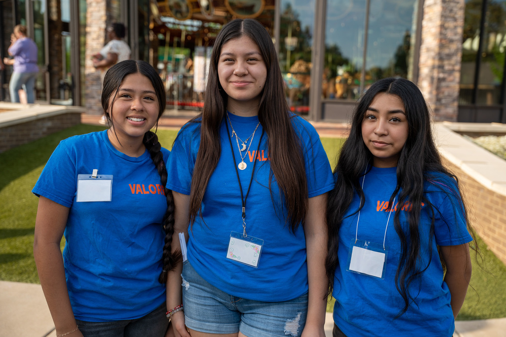 Three female ABSS students representing the Valores organization smile for a photo at Burlington Carousel Park. They are wearing matching bright blue T-shirts with "VALORES" printed in orange.