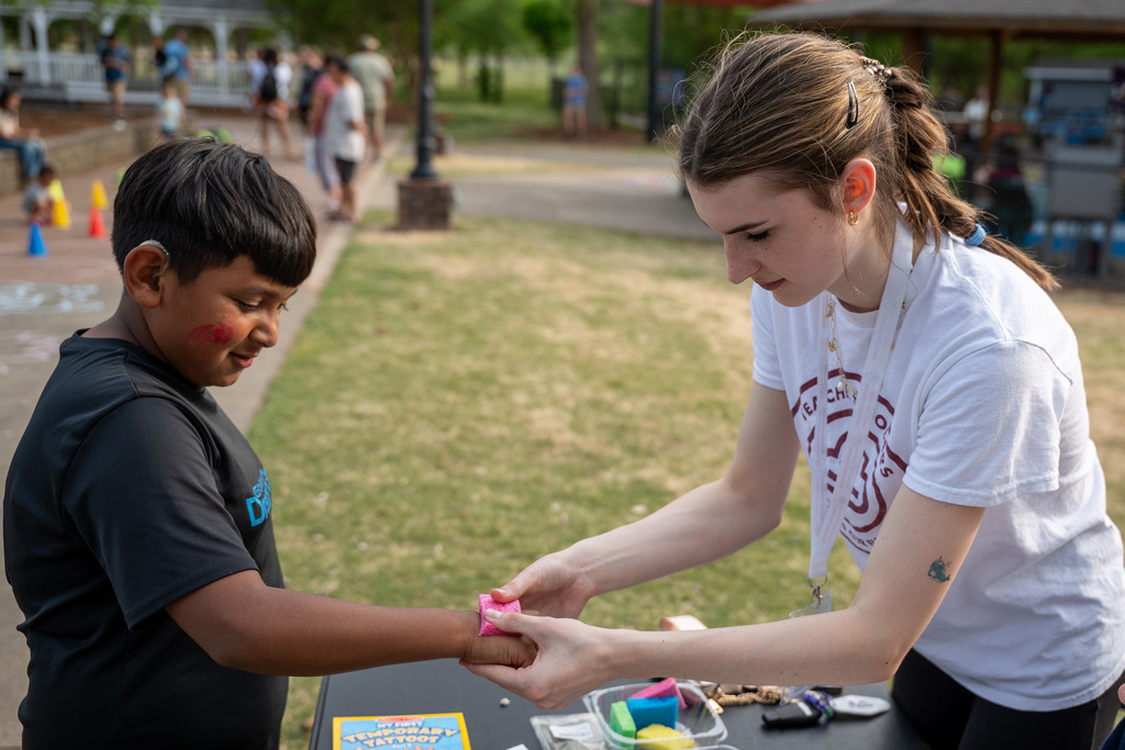 An ABSS Teacher Scholar in a white logo T-shirt carefully applies a temporary tattoo to the arm of a young boy at a sunny outdoor activity station.