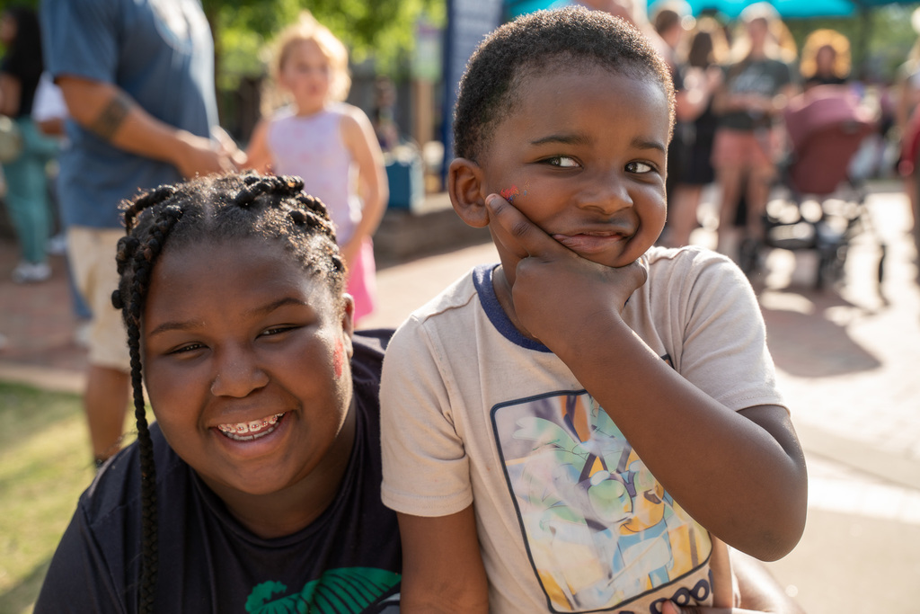 Close-up of two young children smiling at the event; a young boy rests his chin on his hand thoughtfully while a girl with braided hair smiles broadly next to him.