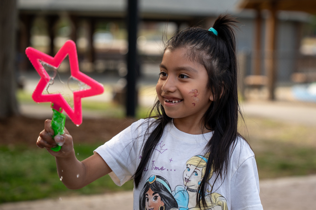 A young girl with a mermaid design painted on her cheek smiles joyfully while holding a star-shaped bubble wand.
