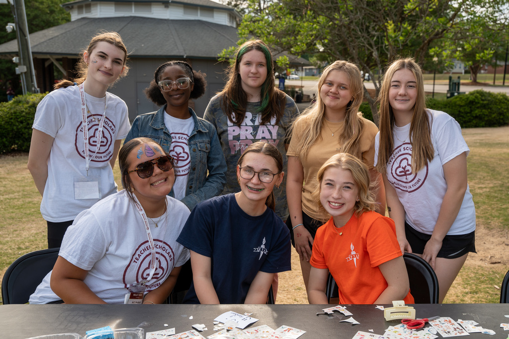 A group of eight ABSS Teacher Scholars smile behind a table. Several are wearing white T-shirts with the Teacher Scholars logo, and some have festive face paint.