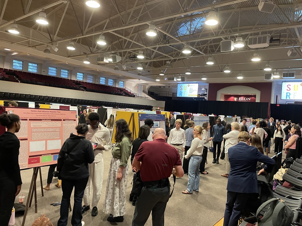 Onlookers walk by and discuss the research projects students are presenting in an Elon gynasium