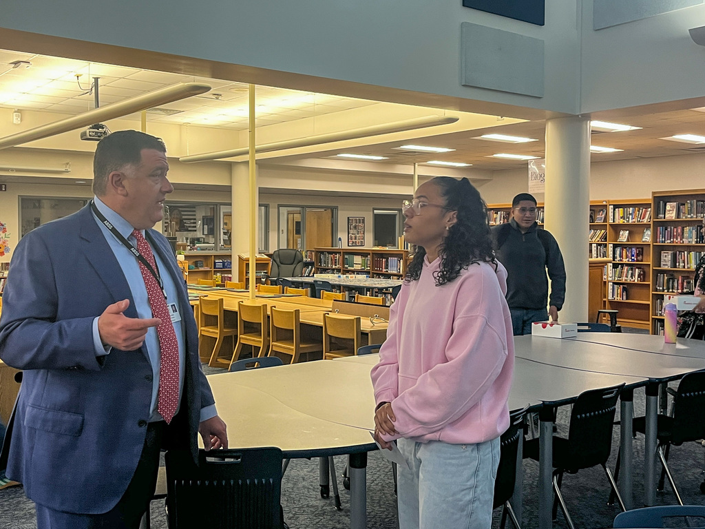 Dr. Fleming, wearing a blue suit and red patterned tie, stands in a library and speaks with a female student wearing a pink hoodie and glasses.