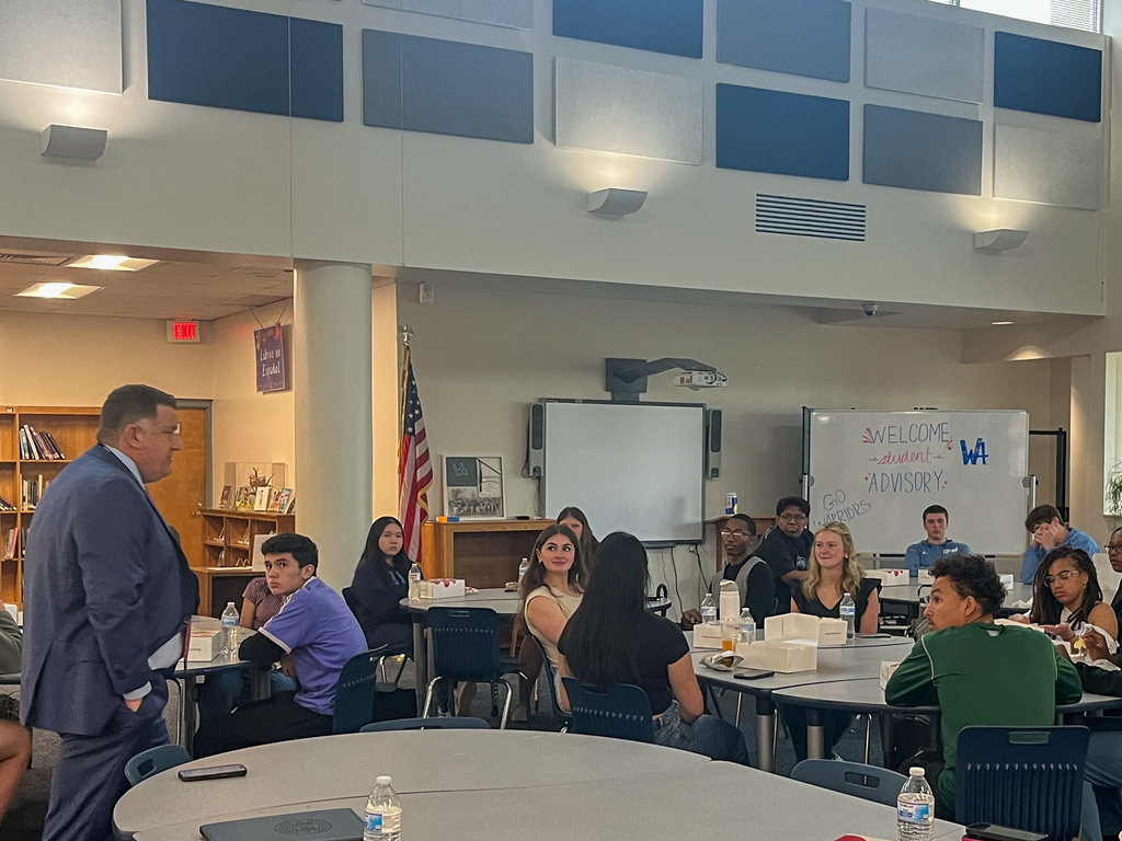 Dr. Fleming stands at the front of a school media center addressing a group of high school students seated at round tables. A whiteboard in the background reads "Welcome Student Advisory" with "Go Warriors" written on the side.