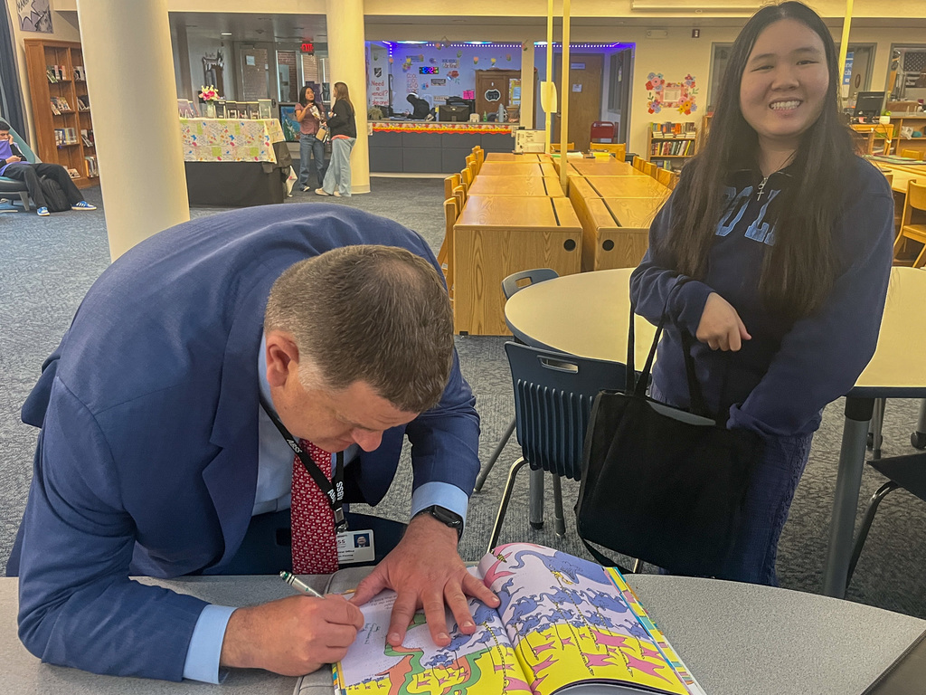 Dr. Fleming leans over a table to write a message in a colorful Dr. Seuss book, "Oh, the Places You'll Go!" as a smiling student in a blue sweatshirt watches.