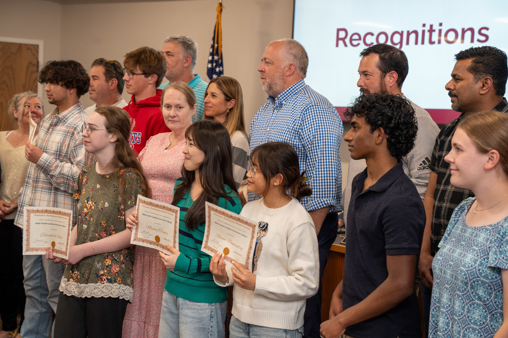 A medium close-up shot of a diverse group of students and supportive adults during a recognition ceremony. Several students in the foreground are smiling and holding certificates for the North Carolina Governor’s School. The atmosphere is celebratory, with a "Recognitions" slide visible on a screen in the background.