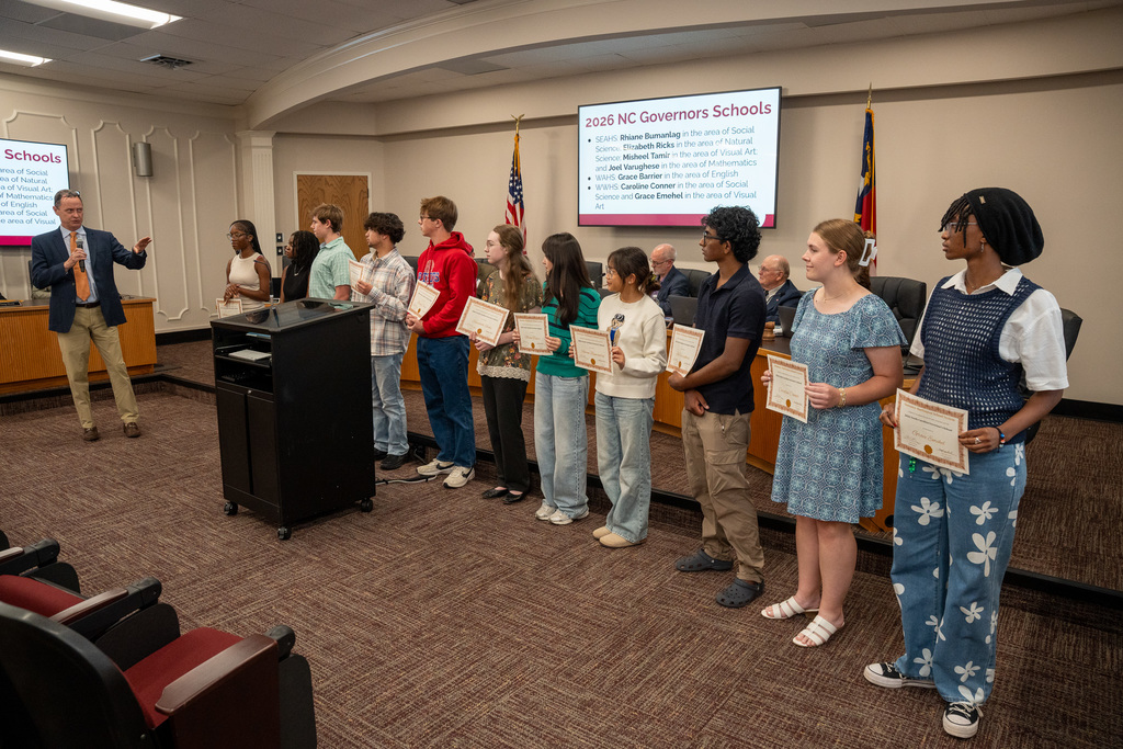 A school board official speaks at a podium to a row of ten high school students standing in a meeting room. The students are holding award certificates and facing the official. A large screen in the background displays a slide titled "2026 NC Governors Schools" with a list of student names and their academic areas of study.
